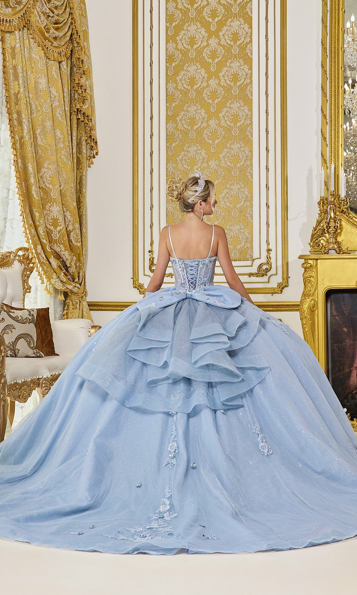 A woman wearing the Quinceanera Dress 5073 By Dancing Queen, featuring a light blue sheer corset bodice, stands with her back to the camera in an ornate gold-accented room with decorative curtains and a fireplace.